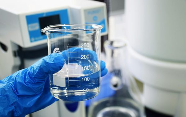 Gloved hand holding beaker of clear PAC solution for water treatment lab test